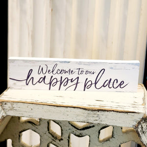 This mini sign has a whitewashed wooden background with hints of greys and turquoise. The words "Welcome to our happy place" are in a dark grey cursive font.   Wood Depth: 0.4375" Width: 6" Height: 1.5"
