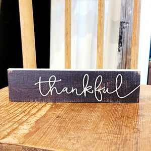 This mini sign has a distressed black background with "thankful" written in a white cursive font.   Wood Depth: 0.4375" Width: 6" Height: 1.5"