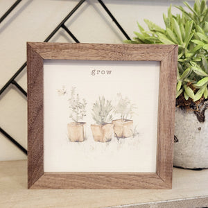A pretty picture of three square terracotta pots with herbs in them. It has the word "grow" above and a butterfly flying on the left.  This unique piece is a simple way to bring beauty and charm to any wall or shelf within the home. It is made from high-quality American hardwood planks with a hand-painted face, printed with UV-cured ink, and is framed in a natural walnut frame. 