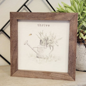 A pretty picture of a watering can with green leaves inside and the word "thrive" above with a bumblebee flying to the left of the picture.  This unique piece is a simple way to bring beauty and charm to any wall or shelf within the home. It is made from high-quality American hardwood planks with a hand-painted face, printed with UV-cured ink, and is framed in a natural walnut frame. 