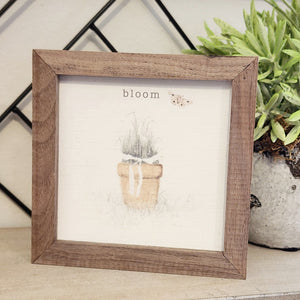 A pretty picture of a pot with flowers tied by a white ribbon.  Above is the word "bloom," with a butterfly off to the right.  This unique piece is a simple way to bring beauty and charm to any wall or shelf within the home. It is made from high-quality American hardwood planks with a hand-painted face, printed with UV-cured ink, and is framed in a natural walnut frame. 