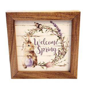 A little bunny holding a purple egg has the word "Welcome Spring" in the center of the picture with a wreath in the center with a pink egg, two butterflies, greens, and lavender going around the wreath.  This unique piece is a simple way to bring beauty and charm to any wall or shelf within the home. It is made from high-quality American hardwood planks with a hand-painted face, printed with UV-cured ink, and is framed in a natural walnut frame.