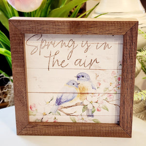 Two bluebirds sit on a blooming dogwood branch with the words "Spring is in the air" above.  This unique piece is a simple way to bring beauty and charm to any wall or shelf within the home. It is made from high-quality American hardwood planks with a hand-painted face, printed with UV-cured ink, and is framed in a natural walnut frame.