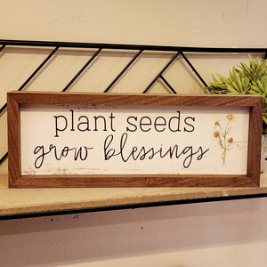 "Plant Seeds, Grow Blessings" is written on a whitewashed background in mixed fonts.  Along the righthand side is a bunch of four daisies.  This unique piece is a simple way to bring beauty and charm to any wall or shelf within the home. It is made from high-quality American hardwood planks with a hand-painted face, printed with UV-cured ink, and is framed in a natural walnut frame.
