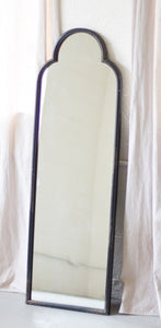 A tall narrow mirror with a thing antiqued black finish irorn frame that has a rounded arch at the top.