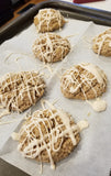 A cookie sheet with parchment paper on it with six round scones drizzled with white frosting on it