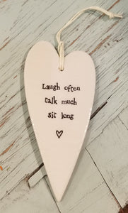 a porcelain white heart shaped ornament with a white cord to hang with the words "Laugh often talk much sit long"