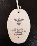 A white oval porcelain ornament with ridges going horizontally.  On the ornament is a line drawing of a bee and underneath is "In a world where you can be anything be yourself" *  in a black typeset font