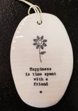 A white oval porcelain ornament with ridges going horizontally.  On the ornament is a line drawing of a flower and underneath is "Happiness is time spent with a friend" *  in a black typeset font