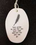 A white oval porcelain ornament with ridges going horizontally.  On the ornament is a line drawing of a feather and underneath is "Take pride in how far you have come and how far you can go" *  in a black typeset font