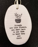 A white oval porcelain ornament with ridges going horizontally.  On the ornament is a line drawing of a potted plant and underneath is "Friends are like flowers, you don't have to see them to know they are there" *  in a black typeset font