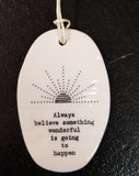A white oval porcelain ornament with ridges going horizontally.  On the ornament is a line drawing of a sunrise and underneath is "Always believe something wonderful is going to happen" *  in a black typeset font