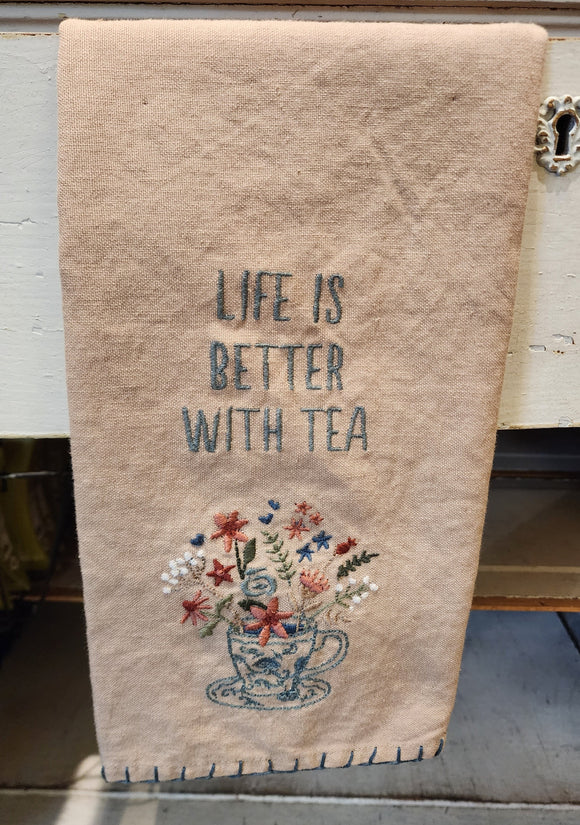 This sweet little tan-colored tea towel has an embroidered teacup in blue on the front with flowers in shades of pink, green, and white along with green leaves, all springing out of it.  Above it says, 
