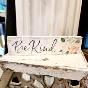 This mini sign has a white-washed wooden background design and the words "Be Kind" in a black cursive font. On the right-hand side are some peach/pink colored flowers with greenery.  Wood Depth: 0.4375" Width: 6" Height: 1.5"