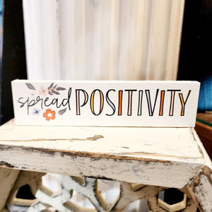 This mini sign has a white background with the words "Spread Positivity" in a black mixed font (with the word "positivity" in pink, yellow, blue and orange letters). Above and below the word "spread" is a cluster of flowers in orange, blue and yellow.  Wood  Depth: 0.4375" Width: 6" Height: 1.5"