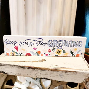 This mini sign has a white background with a mix of wildflowers along the bottom.  Above the flowers it says "Keep going, keep GROWING" in a black mixed font (with the word "growing" in blue).  Wood  Depth: 0.4375" Width: 6" Height: 1.5"