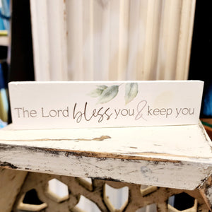 This white mini sign has the words "The Lord bless you & keep you" in a black mixed font.  Above the words "bless" and "you" is a bunch of leaves hanging down.  Wood Depth: 0.4375" Width: 6" Height: 1.5"