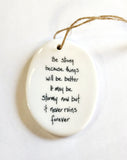 We love our porcelain ornaments because they are perfect for gift-giving! This oval ornament says, "Be strong because things will be better.  It may be stormy now, but it never rains forever" on one side. On the other side is a grey background with a cloud over the water with a sailboat in white.   Dimensions: 2" W x 2 3/4" H
