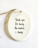 We love our porcelain ornaments because they are perfect for gift-giving! This oval ornament says, "Thank you for being the reason I smile" on one side. The other side has a grey background with a bird sitting on a branch with four birds in the sky, all in white.  Dimensions: 2" W x 2 3/4" H