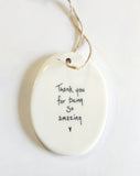 We love our porcelain ornaments because they are perfect for gift-giving! This oval ornament says, "Thank you for being so amazing," with a heart beneath on one side. The other side has a grey background with a branch running the center with flowers all in white.  Dimensions: 2" W x 2 3/4" H