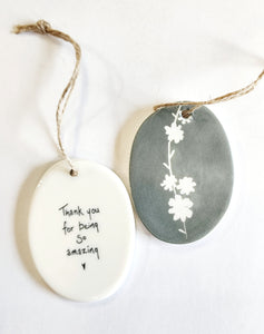 We love our porcelain ornaments because they are perfect for gift-giving! This oval ornament says, "Thank you for being so amazing," with a heart beneath on one side. The other side has a grey background with a branch running the center with flowers all in white.  Dimensions: 2" W x 2 3/4" H