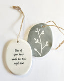 We love our porcelain ornaments because they are perfect for gift-giving! This oval ornament says, "One of your hugs would be nice right now" on one side. The other side has a grey background with a single branch with leaves in white.  Dimensions: 2" W x 2 3/4" H